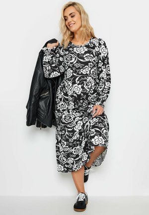 Robe midi florale noir et blanc avec manches longues, col rond et fente sur le côté, portée avec une veste en cuir noir et des baskets.