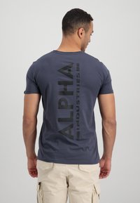 Vyriškas tamsiai mėlynas marškinėliai iš švelnios medvilninės džersio, su dideliu juodu "ALPHA INDUSTRIES" logotipu vertikaliai ant nugaros, apvaliu kaklu ir trumpomis rankovėmis.