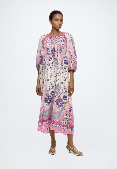Mujer con un vestido midi suelto, floral en tonos rosa y púrpura con mangas abullonadas y sandalias blancas de tacón bajo, de pie contra un fondo liso.