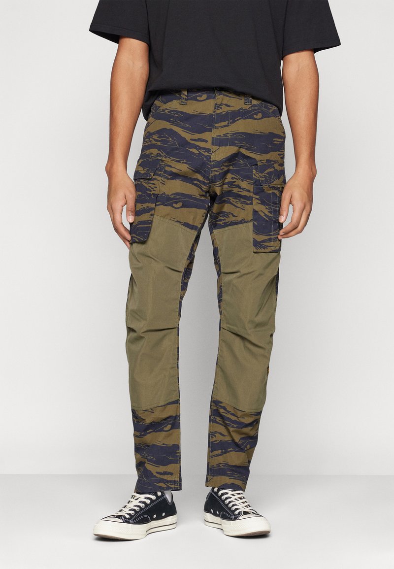 Pantalones cargo en verde oliva con un patrón de camuflaje azul-negro. Presentan bolsillos laterales, un diseño ajustado y tejido resistente. Combinados con zapatillas negras.