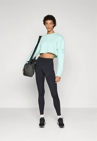 Lichtblauw cropped sweatshirt met lange mouwen en logodetails, gecombineerd met strakke zwarte leggings en zwarte sportschoenen. Zwarte sporttas over de schouder.