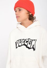 Witte fleece hoodie met een groot zwart "VOLCOM" logo aan de voorkant, gecombineerd met een helder rood geribbeld brei-sjaal.