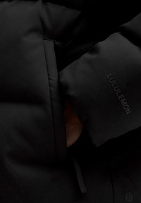 Čierna zateplená bunda s hladkou textúrou, viditeľným šitím, vreckom na zips a logom Lululemon vytlačeným písmenami.