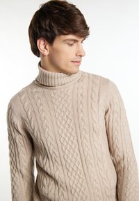 Beige színű cable-knit garbó pulóver, texturált mintával, vastag bordázással a gallérnál és bonyolult díszítőelemekkel a felületen.