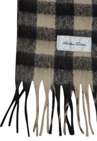 Rutet tørklæde med skiftevis sorte og beige firkanter, frynsekanter, lavet af blødt materiale. Har et hvidt mærke med "Another Cotton" i sort.