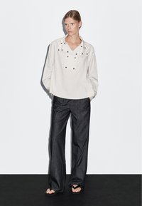 Pruhovaná, oversized světle béžová košile s širokým límcem a černými patentkami, doplněná volnými tmavými džínovými kalhotami a plochými sandály.