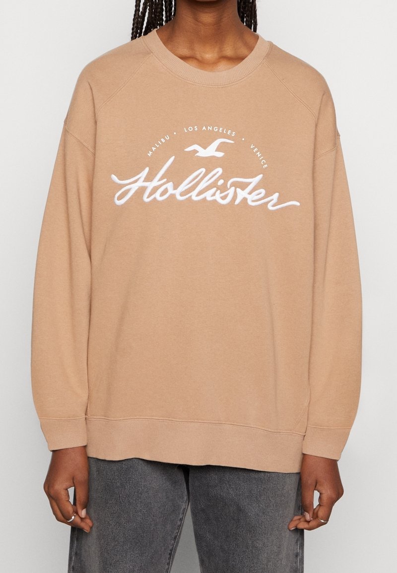 Beige sweatshirt med rund halsringning, långa ärmar och ribbade muddar. Har en vit broderad "Hollister"-logotyp och grafisk text.