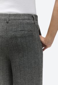 Približni posnetek osebe, ki nosi sive hlače z vzorcem ribje kosti, z zadnjim žepom z odprtino, roka počiva na boku, bela majica je speta notri.