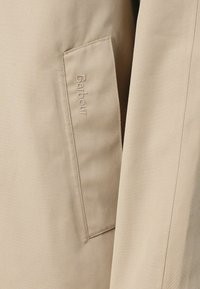 Tecido leve em bege com um bolso frontal apresentando o logótipo "Barbour" em relevo. Textura suave, design ajustado, estilo minimalista.