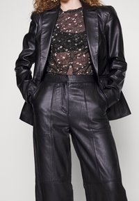 Zwarte leren blazer over een zwarte doorzichtige bloes met bloemenprint, gecombineerd met hooggerokte zwarte leren wijde broek met paneelstiksels.