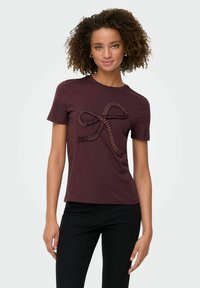 Kobieta z kręconymi włosami, ubrana w ciemnoczerwoną koszulkę z teksturowanym wzorem kokardy oraz czarne spodnie, stojąca na jednolitym tle.