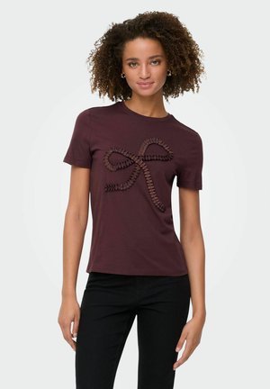 Kvinde med krøllet hår iført en mørk burgunder t-shirt med et tekstureret sløjfedesign og sorte bukser, stående mod en ensfarvet baggrund.