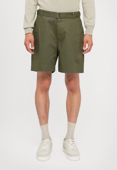 Groene katoenen cargoshorts met twee grote zijzakken, een riem met metalen gesp en een rechte snit. Draagt met beige sokken en sneakers.