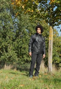 Sort puffejakke med lynlås foran, stående krave og struktureret overflade, parret med sorte bukser og mørke fodtøj i et grønt udendørs miljø.