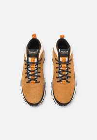 Zapatillas de ante color beige con suela negra y blanca. Cuentan con cordones negros, acentos naranjas y un cuello acolchado. Logo de Timberland en la lengüeta.