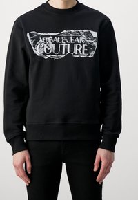 Svart sweatshirt av mjukt tyg, med ett slitet grafiskt tryck över bröstet med texten "Versace Jeans Couture" i vitt.