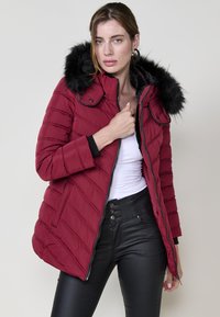 Sarkans pūkains mētelis ar noņemamu melnu kažokādas odere, quilted dizains, ar rāvējslēdzēju aizdari un pieguļošu aproču. Nēsāts pār balto augšu.