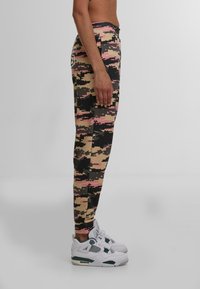 Leggings s kamuflažnim uzorkom u bež, crnoj i ružičastoj boji sa pripijenim dizajnom. Upareni s bijelim tenisicama s zelenim detaljima i mrežastim elementima.