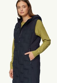 Weste mit Kapuze in Dunkelblau, ausgestattet mit einem gesteppten Muster und seitlichen Taschen, über einem grünen Langarmshirt getragen.