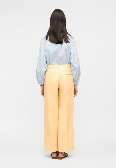 Femme debout de dos, portant une blouse bleu clair avec de petites étoiles brodées et un pantalon large taille haute jaune, accompagnée de sandales.