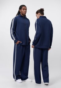Navy athletic tracksuit featuring a zip-up jacket and wide-leg pants, both with white side stripes and pockets. Made from soft fabric.