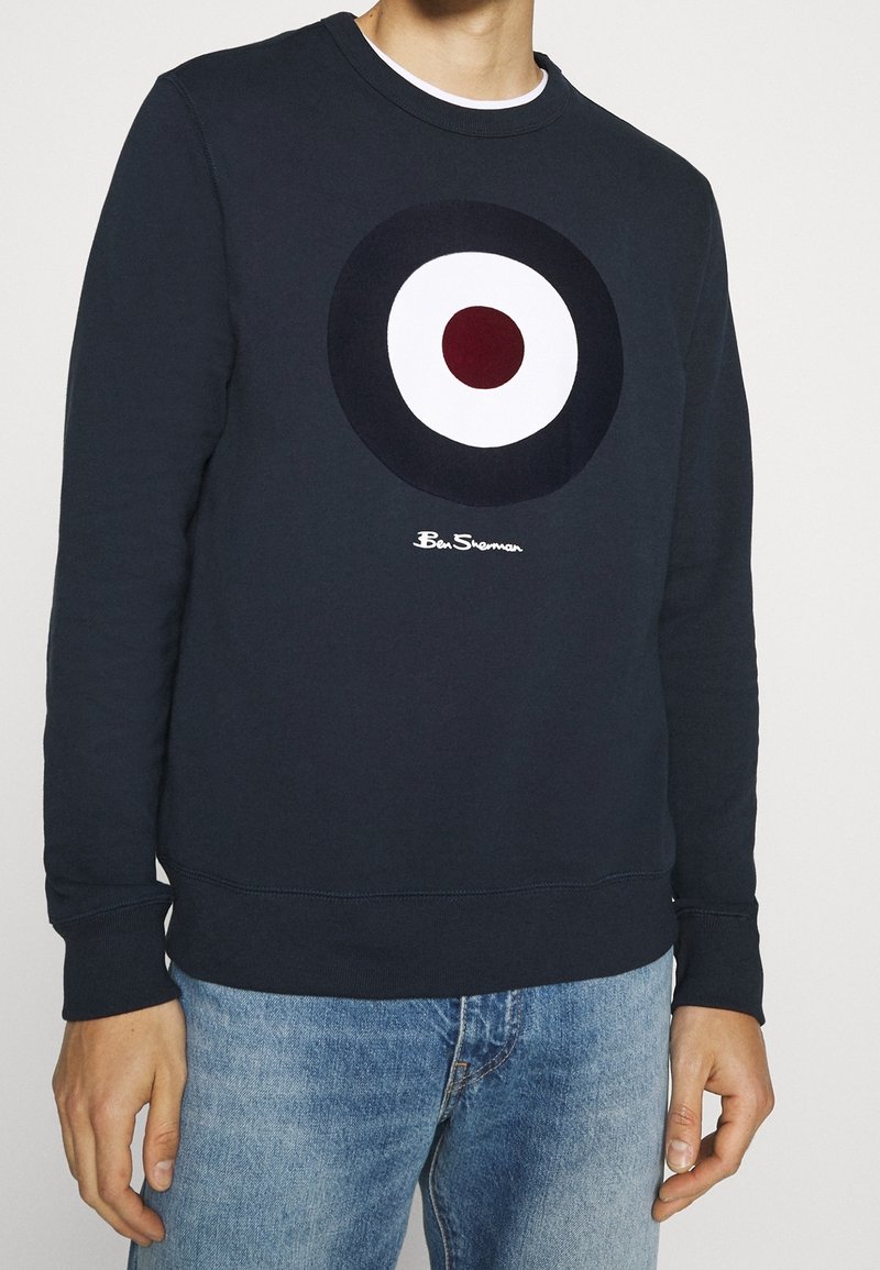 Homme portant un sweat-shirt bleu marine avec un motif de cible à cercles concentriques et le logo "Ben Sherman", associé à un jean bleu clair.