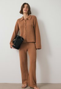 Pleteni rjavi dvodelni set, ki vsebuje z gumbi zapenjanje kardigan z ovratnikom in široke hlače. Tekstura je odprto tkana, kroj pa je ohlapen.
