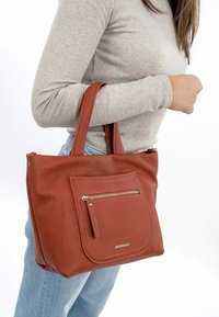 Borsa in pelle rossa con texture, dal design spazioso, caratterizzata da una tasca frontale con zip e doppi manici, tenuta da una persona in jeans azzurri chiari.