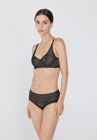 Černá krajková souprava spodního prádla obsahující braletku s nastavitelnými ramínky a vlnitými okraji, doplněná o odpovídající kalhotky s nízkým pasem.
