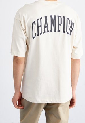 Persona che indossa una maglietta larga color panna con la scritta "CHAMPION" stampata in grandi lettere nere sulla schiena, abbinata a pantaloni beige.