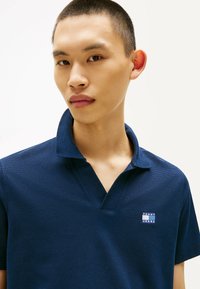 Camisa polo azul marino en tejido texturizado con collar corto, con un pequeño parche del logo de Tommy Jeans en el lado izquierdo del pecho.