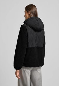 Zwarte hoodie-jack met een gewatteerde bovenkant en een zachte onderkant. Voorzien van elastische manchetten en een ontspannen pasvorm.