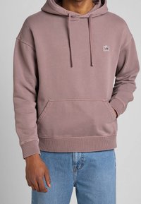 Hooded sweatshirt in soft, dusty rose fabric. Features a front pocket, ribbed cuffs, and a small logo label on the chest. Cotton blend.