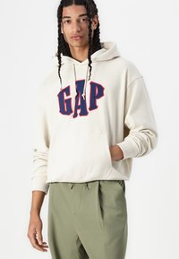 Krémová mikina s kapucňou a logom "GAP" v námorníckej modrej a červenej farbe. Obsahuje šnúrky, klokanie vrecko a ribbed manžety. V kombinácii s olivovými nohavicami.