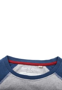 Grå og blå baseball-stil t-shirt med rød detalje ved halsudskæringen. Har en blød tekstur og kontrasterende ærmer. Mærket er synligt indeni.