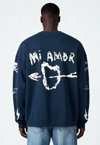 Sweatshirt bleu marine avec un texte graphique blanc "Mi Amor", des cœurs et des motifs de skull. Il présente des poignets et un ourlet côtelés avec des accents en forme d'éclair.