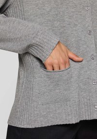Grauer Cardigan mit geripptem Muster, V-Ausschnitt, Knöpfen vorne und Seitentaschen. Der Stoff wirkt weich und warm.