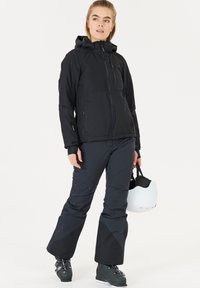 Zwarte geïsoleerde jas met een capuchon, een ritssluiting aan de voorkant en zijzakken; gecombineerd met donkere broek. Model houdt een witte helm vast.