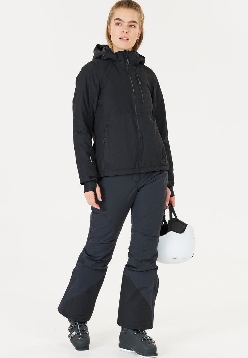 Zwarte geïsoleerde jas met een capuchon, een ritssluiting aan de voorkant en zijzakken; gecombineerd met donkere broek. Model houdt een witte helm vast.