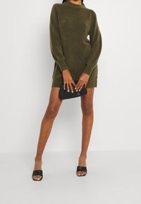 Abito in maglia verde oliva con maniche lunghe, scollatura rotonda e spacchi laterali; abbinato a sandali neri e una pochette nera.