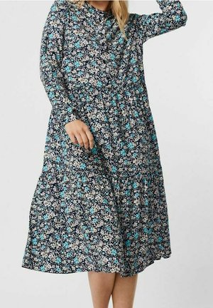 Kobieta ubrana w sukienkę z długim rękawem do kolan w niebieski, biały i czarny kwiatowy wzór, stojąca na jednolitym tle.