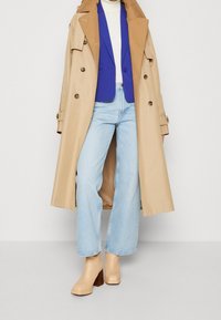 Béžový trenčkot přes modrý blejzr, bílý rolák a světlé modré džíny se širokými nohavicemi. Tělové kotníkové boty na podpatku se čtvercovou špičkou.