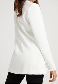 Blazer blanc sur mesure à manches longues, avec une texture lisse, une coupe structurée et trois boutons décoratifs sur les poignets.