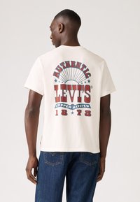 Bijela pamučna majica s retro crveno-plavim grafikonom na leđima s natpisom "AUTENTIČNE LEVI'S® BAKRENE RIVETE 1873." Udoban kroj.