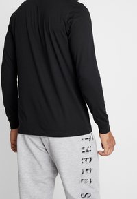 Schwarzes Langarmshirt mit rundem Halsausschnitt, kombiniert mit grauen Jogginghosen, die an der Seite mit einem vertikalen Grafikdruck in dunkler Schrift verziert sind.