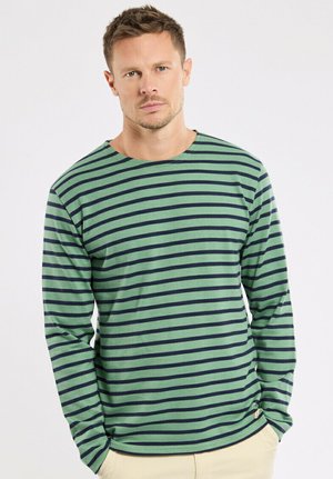 Homme aux cheveux courts portant une chemise à manches longues rayée verte et bleu marine, debout les mains dans les poches sur un fond blanc uni.