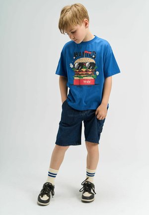 Modrá tričko s krátkym rukávom s grafickým motívom hamburgeru v slnečných okuliaroch. Nosí sa s tmavými džínovými šortkami a čiernymi teniskami s béžovými akcentmi.