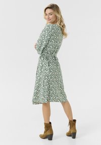 Rochie florală verde cu m�âneci lungi, talie strânsă și fustă vaporoasă. Purtată cu botine din suede maro până la glezne, pe un fundal alb.