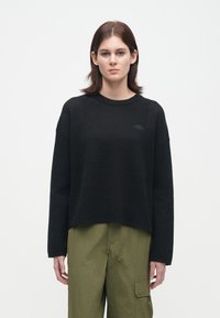 Γυναίκα με καστανά μαλλιά μέχρι τους ώμους, φορώντας ένα μαύρο πουλόβερ μακρύ μανίκι και ελαιόχρωμα cargo παντελόνια, κοιτώντας μπροστά σε άσπρο φόντο.