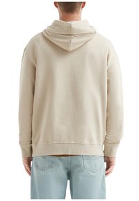 Persona che indossa una felpa beige e jeans azzurri, vista da dietro su uno sfondo bianco.